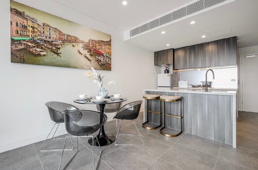 A modern dining area features a round glass table surrounded by four sleek, transparent chairs. An island bench with metallic accents is equipped with high-backed stools. A large, colorful artwork of a canal scene adds a vibrant touch to the light-filled space.