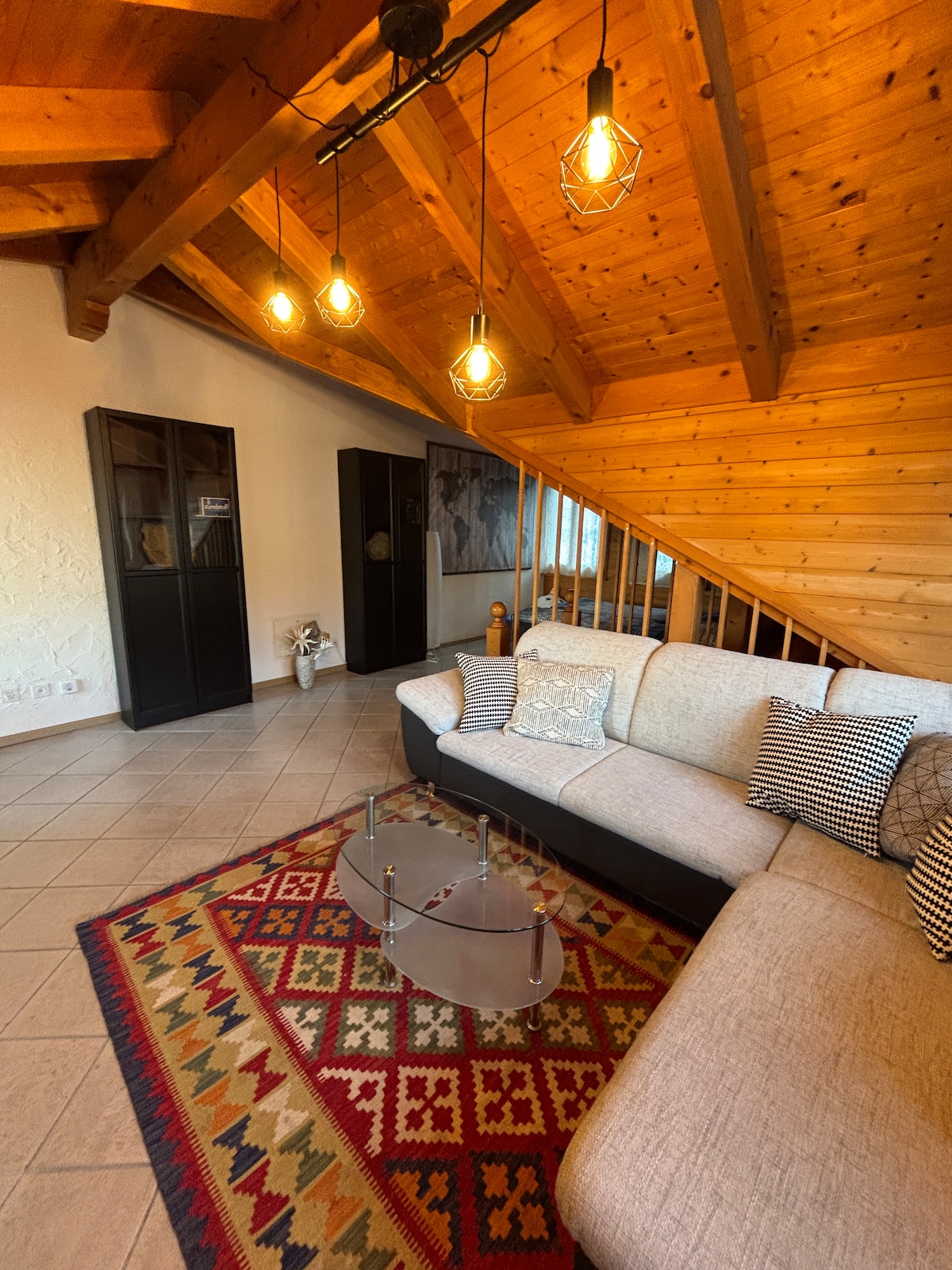 A spacious living area features a cozy sectional sofa adorned with patterned pillows. A glass coffee table sits on a colorful area rug. Warm lighting is provided by industrial-style pendant lights hanging from the wooden ceiling beams, creating an inviting ambiance.