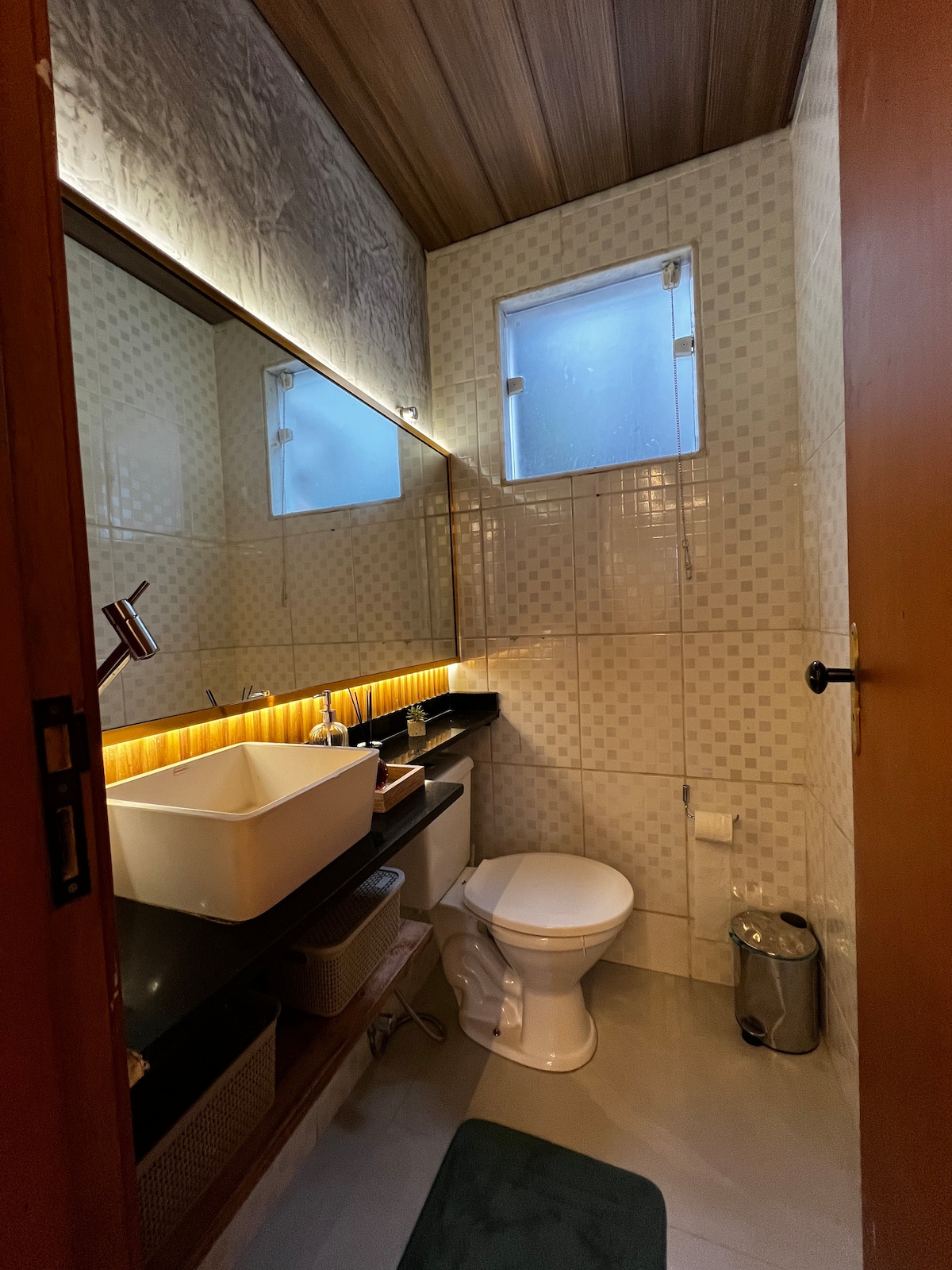 The bathroom features a modern design with a black countertop and a well-lit mirror. A white sink and a toilet are situated against tiled walls. Natural light enters through small windows, and a dark green mat adds a touch of color to the gray floor.