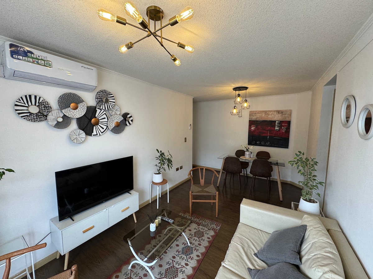 A modern living area features a comfortable cream-colored sofa and a sleek television stand. Above, decorative wall pieces add visual interest. A dining area with dark chairs and a round table is visible, alongside green plants that enhance the space's inviting feel.