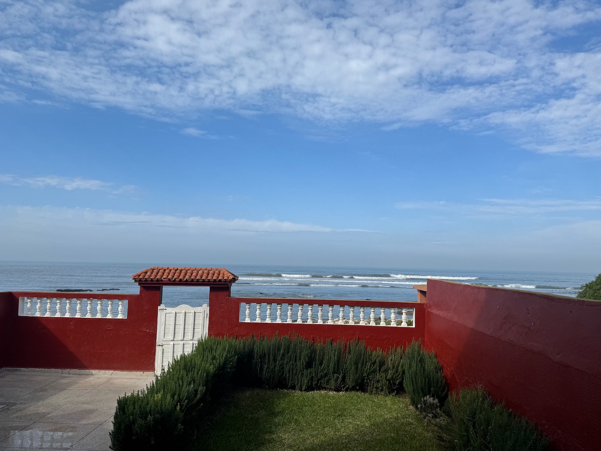 A scenic outdoor area features a red wall with a white gate, offering expansive views of the ocean and waves. Lush greenery surrounds the space, enhancing the peaceful seaside ambiance. The sky is clear with soft clouds, creating a serene atmosphere.