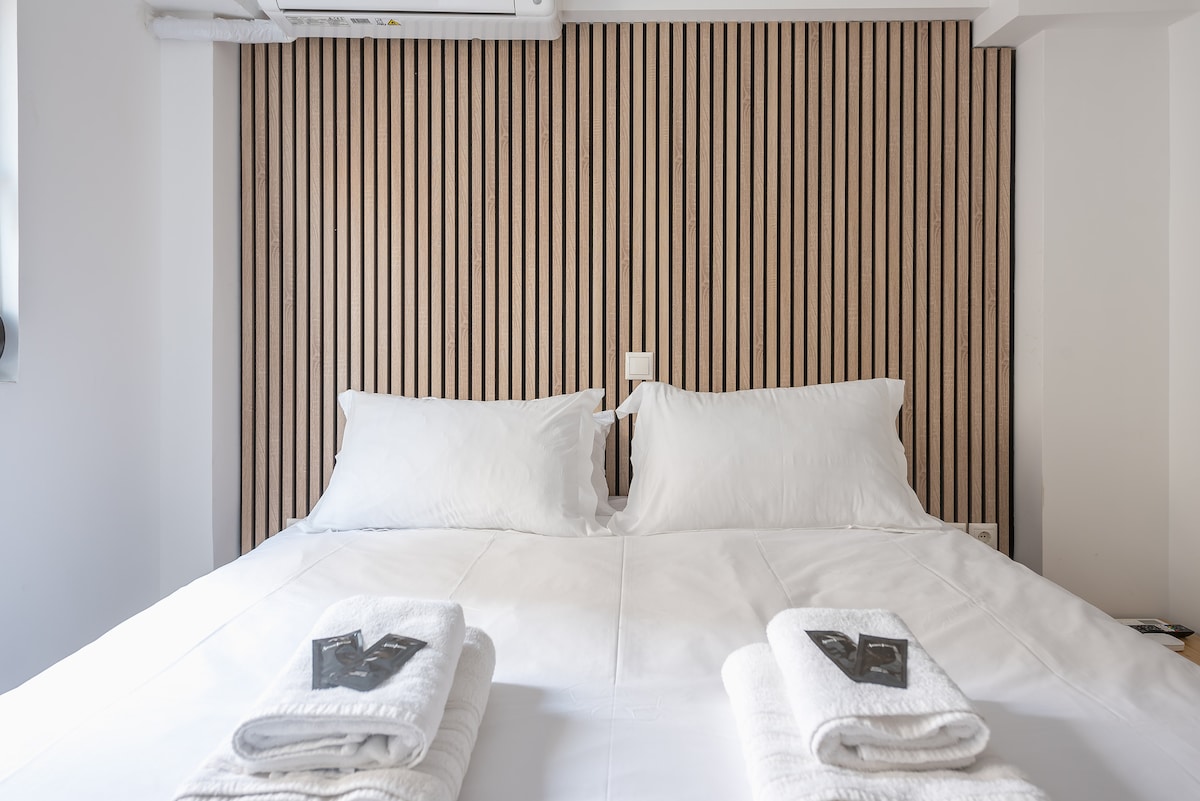 A spacious bed with a white comforter and two fluffy pillows is showcased. The wooden feature wall adds a subtle texture, while neatly folded towels enhance the inviting presentation. Natural light filters in, highlighting the relaxing ambiance of the sleeping area.