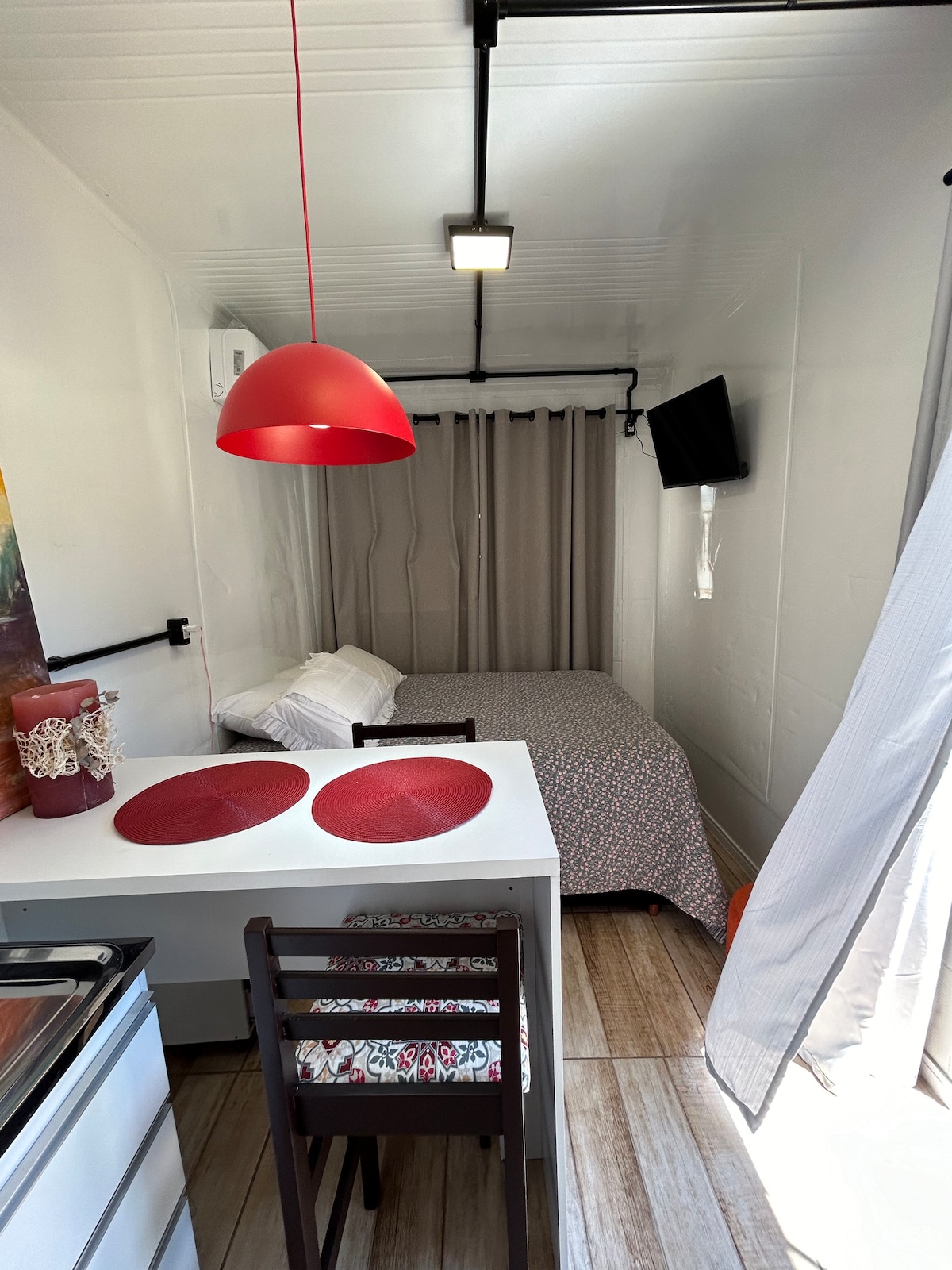 An efficiently designed interior features a dining table with two chairs beside a small kitchenette. A compact bed is situated against the wall, covered in a patterned bedspread. Soft light is provided by a modern pendant light, and a wall-mounted TV is visible.