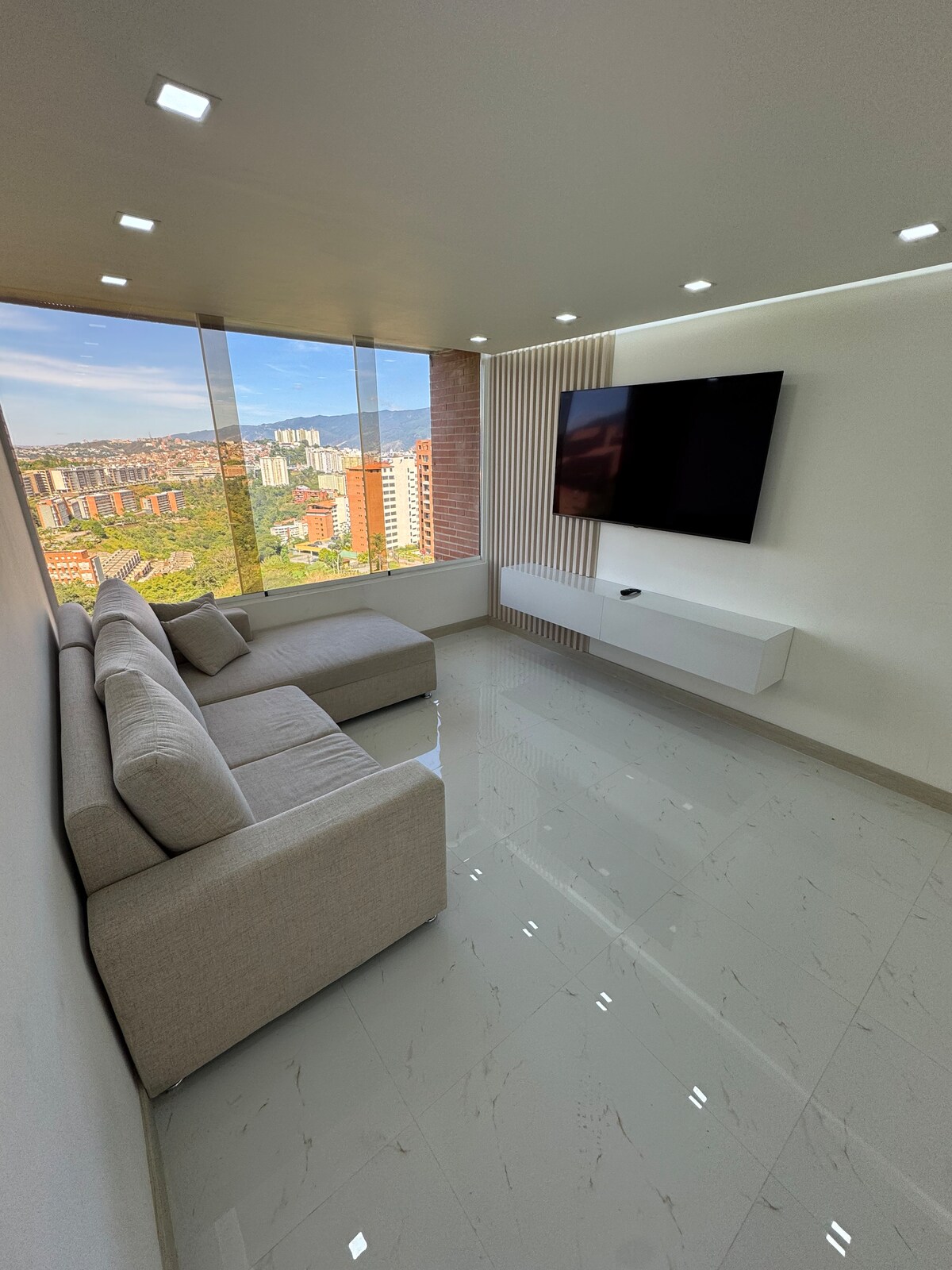 A modern living room features a light-colored, spacious sectional sofa facing a mounted flat-screen television. Floor-to-ceiling windows provide scenic views of the surrounding city and landscape. Bright lighting and glossy tile flooring enhance the open and airy ambiance.