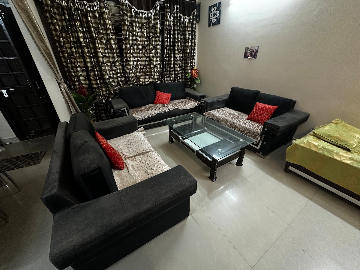 A welcoming living area is furnished with multiple black sofas, accented by red cushions. A glass coffee table sits in the center, complementing the sleek design. Dark, patterned curtains frame the space, while light tiles enhance the room's spacious feel.