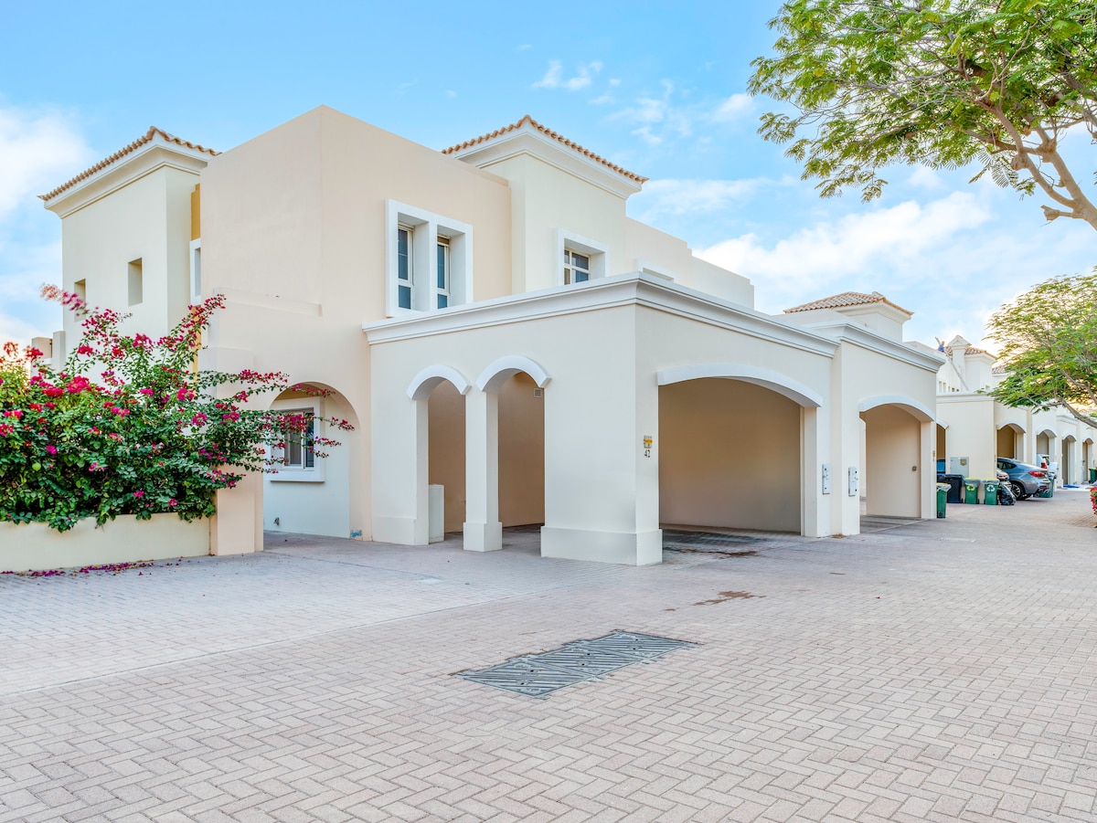 The exterior of the villa features a light-toned façade with arched entrances. Lush greenery and blooming flowers frame the entrance, providing a welcoming atmosphere. A paved walkway leads to the main entrance, while shaded parking spaces are visible in the background.