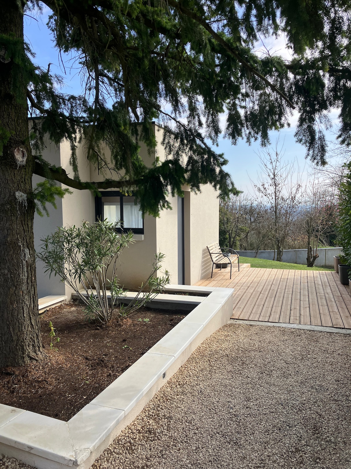 A modern exterior view of the petite maison is visible, framed by a tall conifer tree. A wooden deck extends alongside a landscaped garden, featuring a small, simple water feature surrounded by gravel and plants. A bench provides a seating area, inviting relaxation.