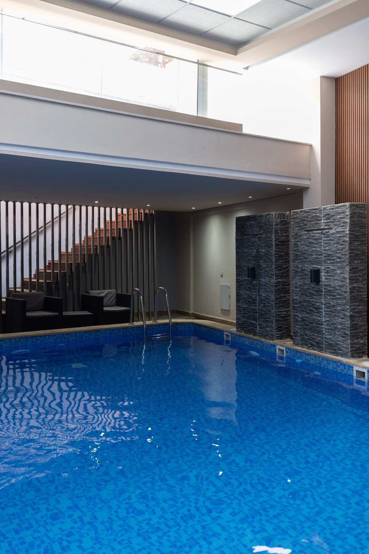 An indoor swimming pool features clear blue water, with stone walls for a modern aesthetic. Smooth steps lead into the pool, and natural light enters through a large window, creating a serene environment. A staircase can be seen in the background, enhancing the architectural design.