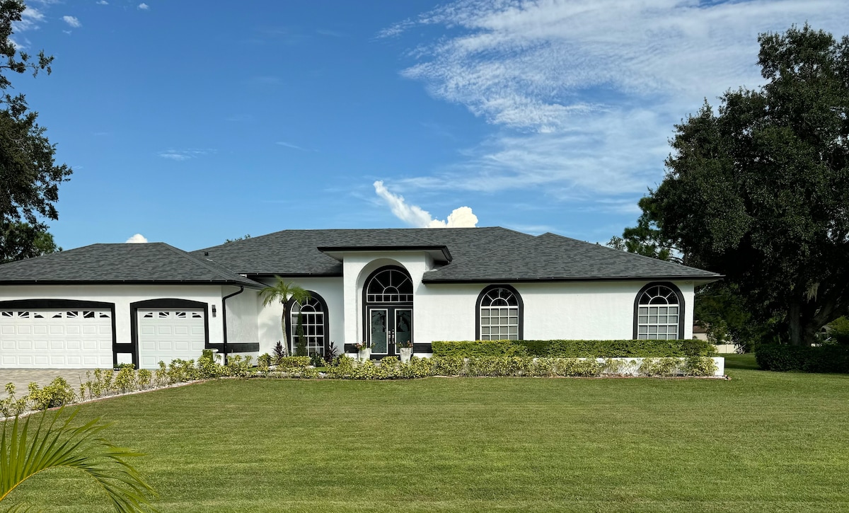 A modern single-story home features a clean white exterior with contrasting black trim. Large arched windows allow natural light to brighten the interior. The well-maintained front yard is covered with lush green grass and bordered by neatly trimmed hedges, creating a welcoming atmosphere.