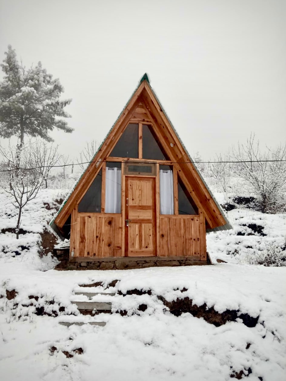 A triangular wooden cabin stands amidst a snowy landscape, featuring a sloped roof and a central door framed by windows. The exterior showcases natural wood finishes, while the ground is covered in a blanket of fresh snow, creating a picturesque winter scene.