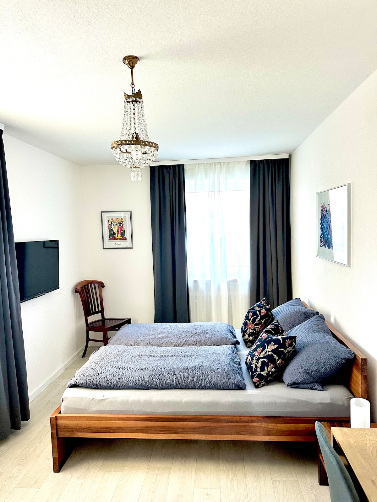 A well-lit bedroom features a spacious bed dressed in soft linens and accented with decorative pillows. A charming chandelier hangs from the ceiling, while a sleek television is mounted on the wall. A wooden chair and a window draped with curtains are positioned nearby.