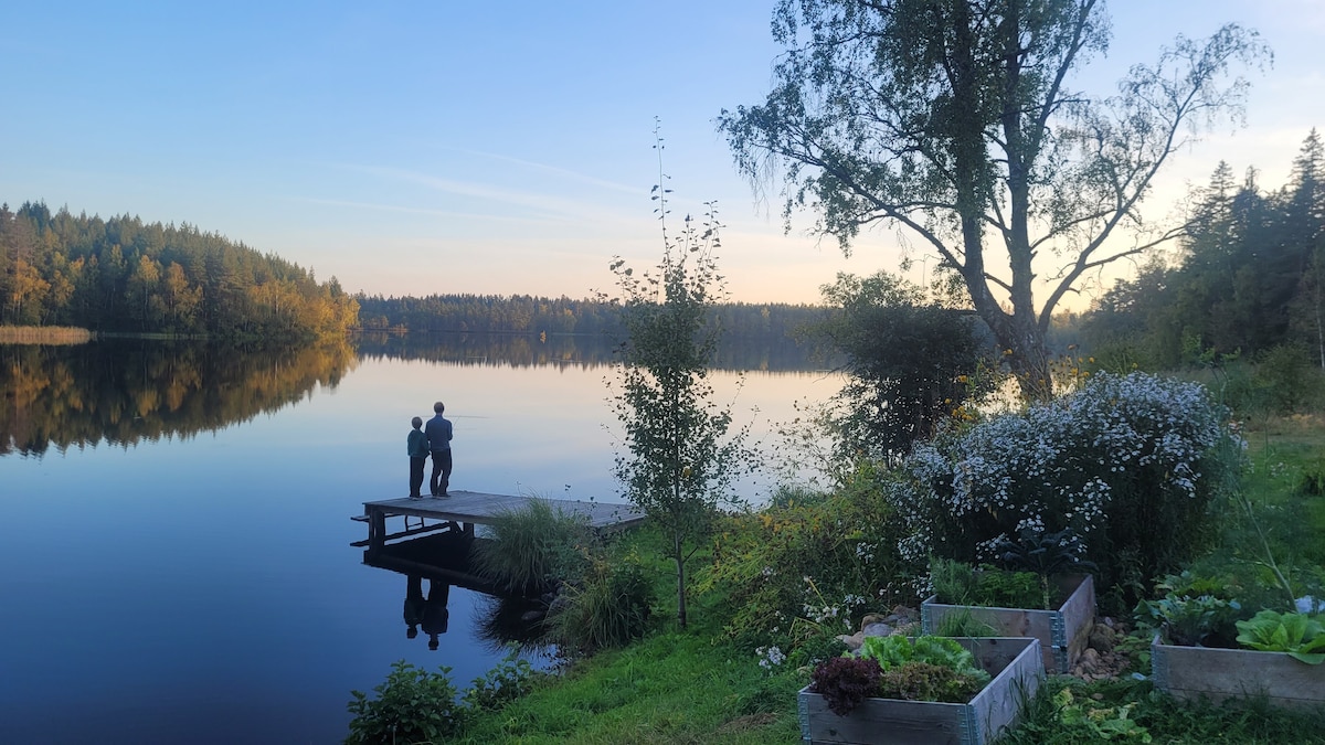 Two figures stand on a wooden dock, silhouetted against a calm lake reflecting the evening sky. Lush greenery and flower beds frame the scene, while trees line the opposite shore, creating a serene backdrop.