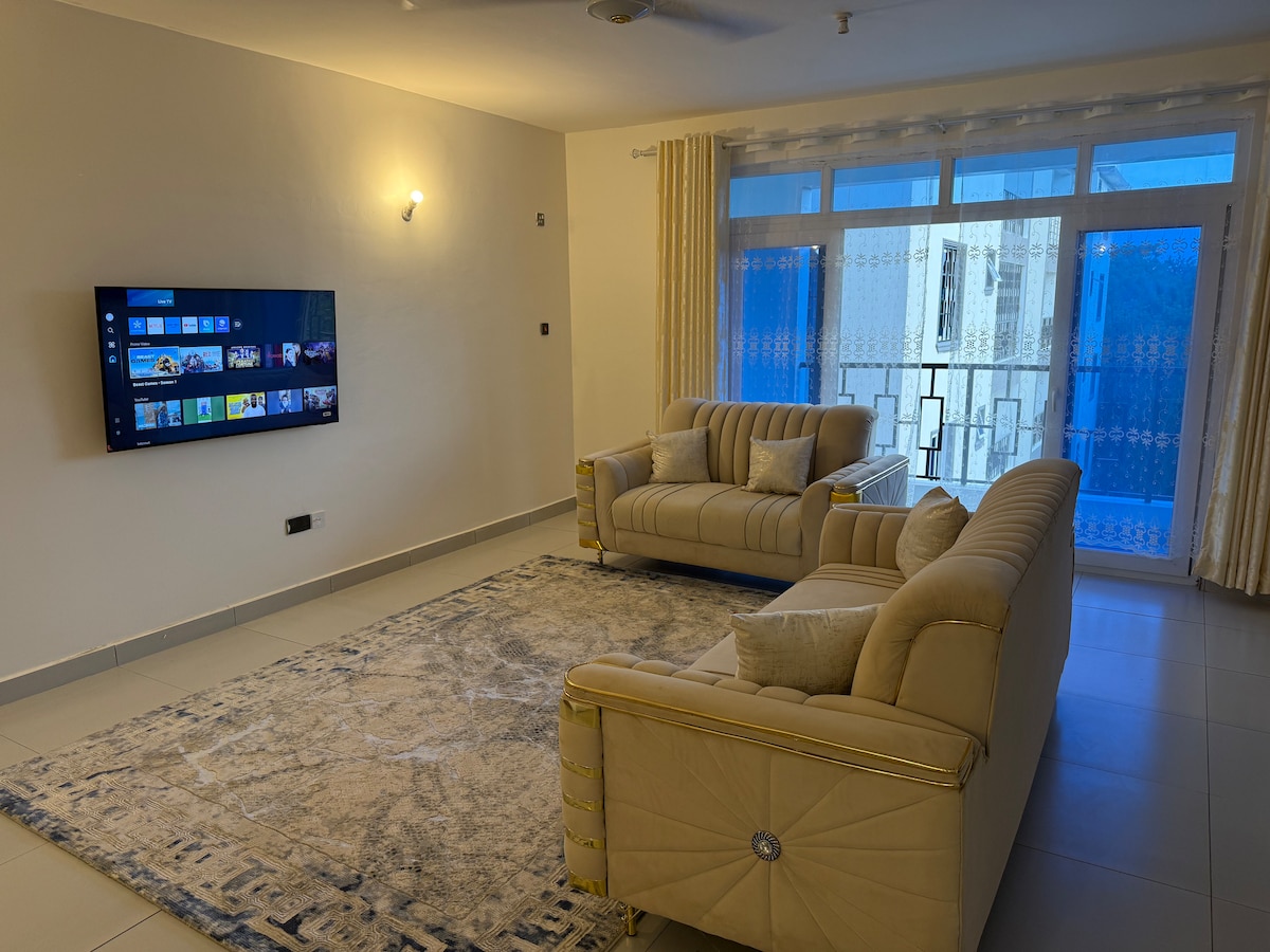 A spacious living area features a large, comfortable sofa set arranged around a soft area rug. A wall-mounted smart TV provides entertainment options, while large windows allow natural light to fill the room, complemented by light-colored curtains.
