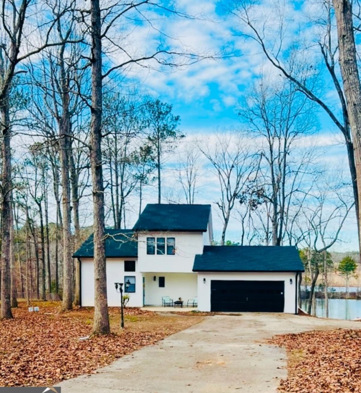 A two-story house with a contemporary design is set among tall trees, featuring a dark roof and white exterior. The driveway leads to a two-car garage, and a small seating area is visible near the front entrance, with a serene lake in the background.