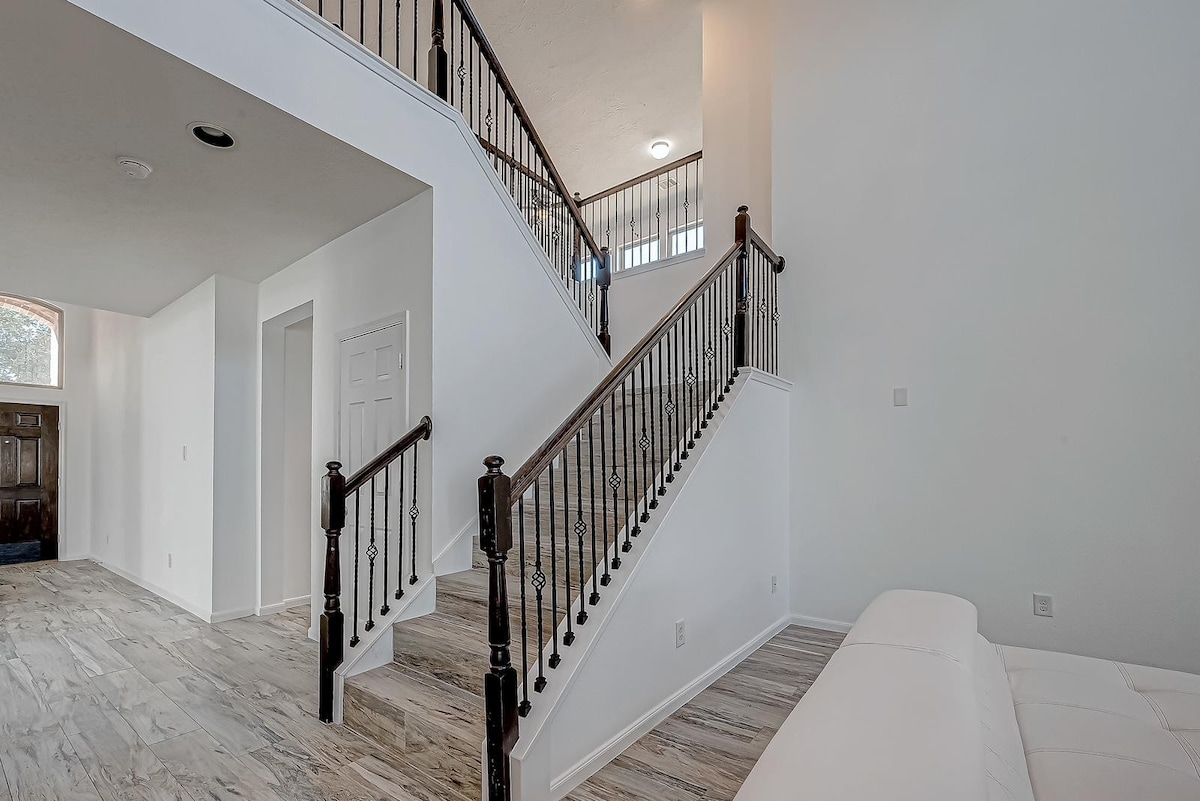 A spacious foyer features an elegant staircase with dark wooden railings leading to the upper level. The light-colored walls and flooring enhance the airy feel of the area. A doorway on the left leads to another room, contributing to the open layout.