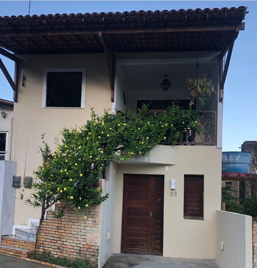 The exterior of a two-story building features a welcoming entrance with a wooden door and a decorative lantern. Lush greenery, including flowering plants, gracefully climbs up the stairs leading to a balcony. The surrounding area shows a mix of natural elements and structured layout.