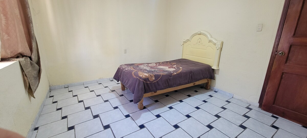 A modest bedroom features a spacious bed with a decorative blanket, positioned against a neutral wall. The tiled floor is designed with alternating light and dark squares. Natural light filters through a partially open window draped with soft curtains. A wooden door adds a touch of warmth.