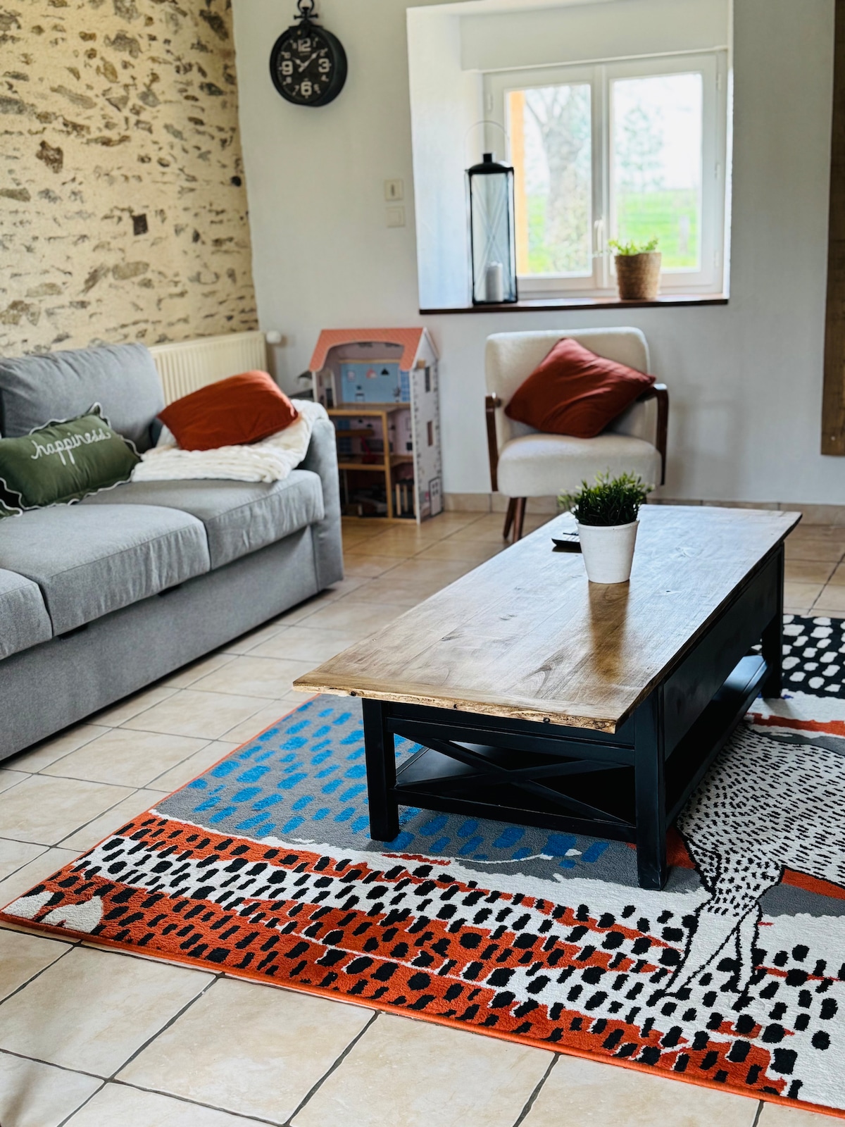 A cozy living area features a soft grey sofa with decorative cushions. A rustic wooden coffee table sits atop a vibrant rug, while a small plant adds a touch of greenery. Natural light enters through a window, illuminating the charming space.