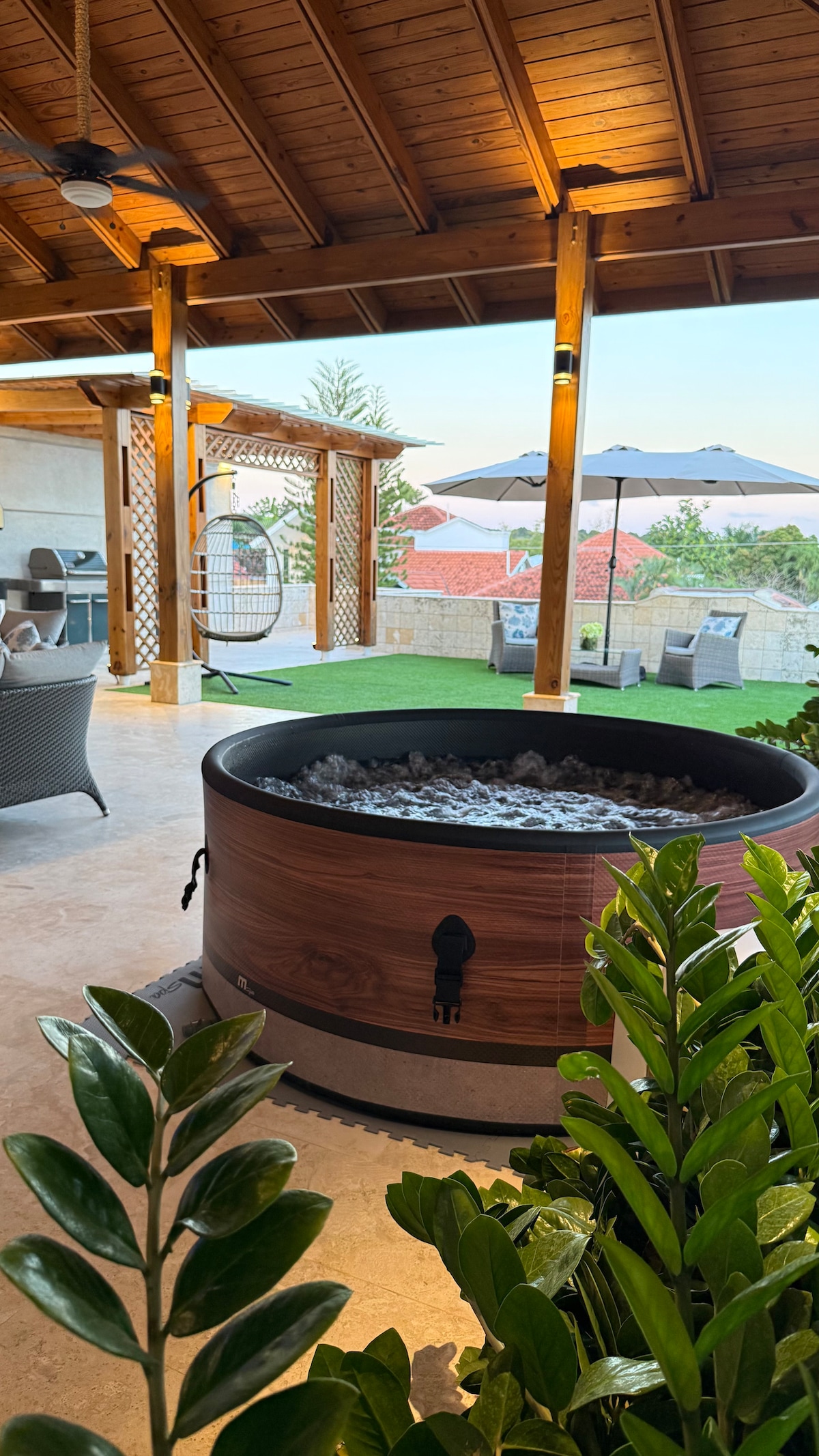 A spacious outdoor area features a wooden hot tub filled with smooth stones. Potted greenery is positioned nearby, while lounge seating is visible in the background. Umbrellas and a scenic view of the surrounding landscape complete the inviting space.
