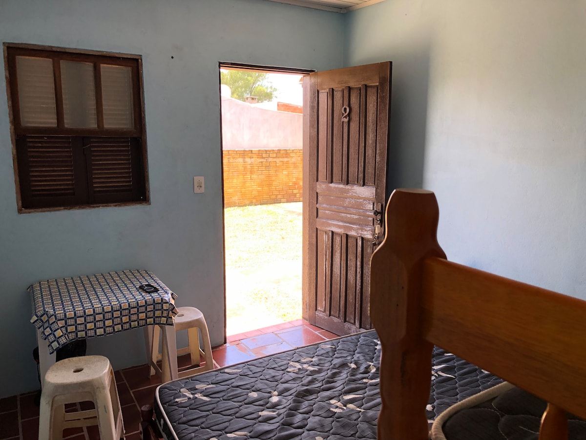 A room with light blue walls features a bed with an unmade mattress and wooden bed frame. A small table covered with a checkered cloth is set against the wall, accompanied by two white stools. The open door reveals a sunny outdoor space.