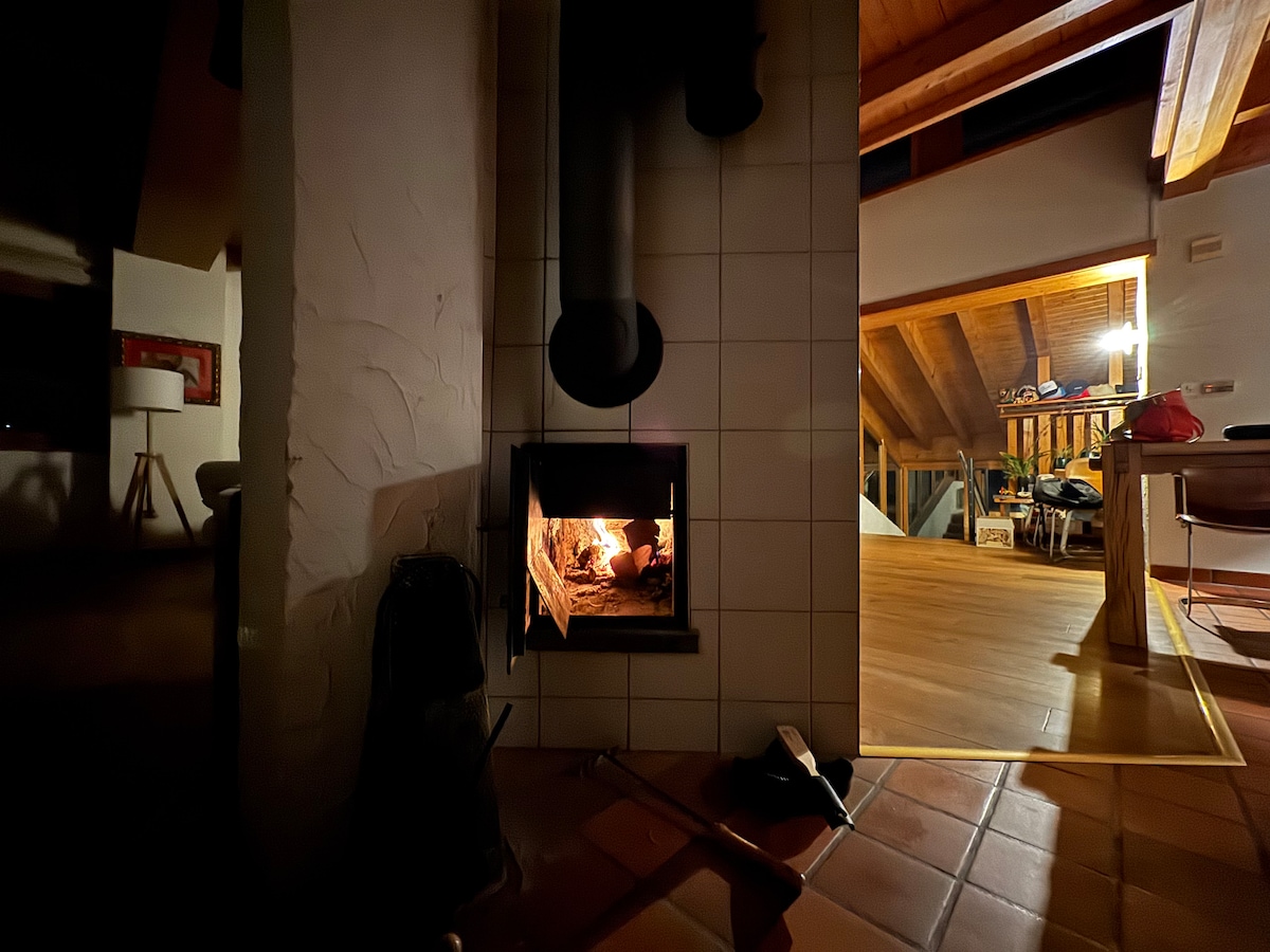 An original 'pierre olaire' fireplace provides warmth, with flames visible in the hearth. The surrounding area reveals wooden beams and a glimpse of the mezzanine floor, emphasizing the home’s spaciousness. Soft, ambient lighting enhances the welcoming environment.