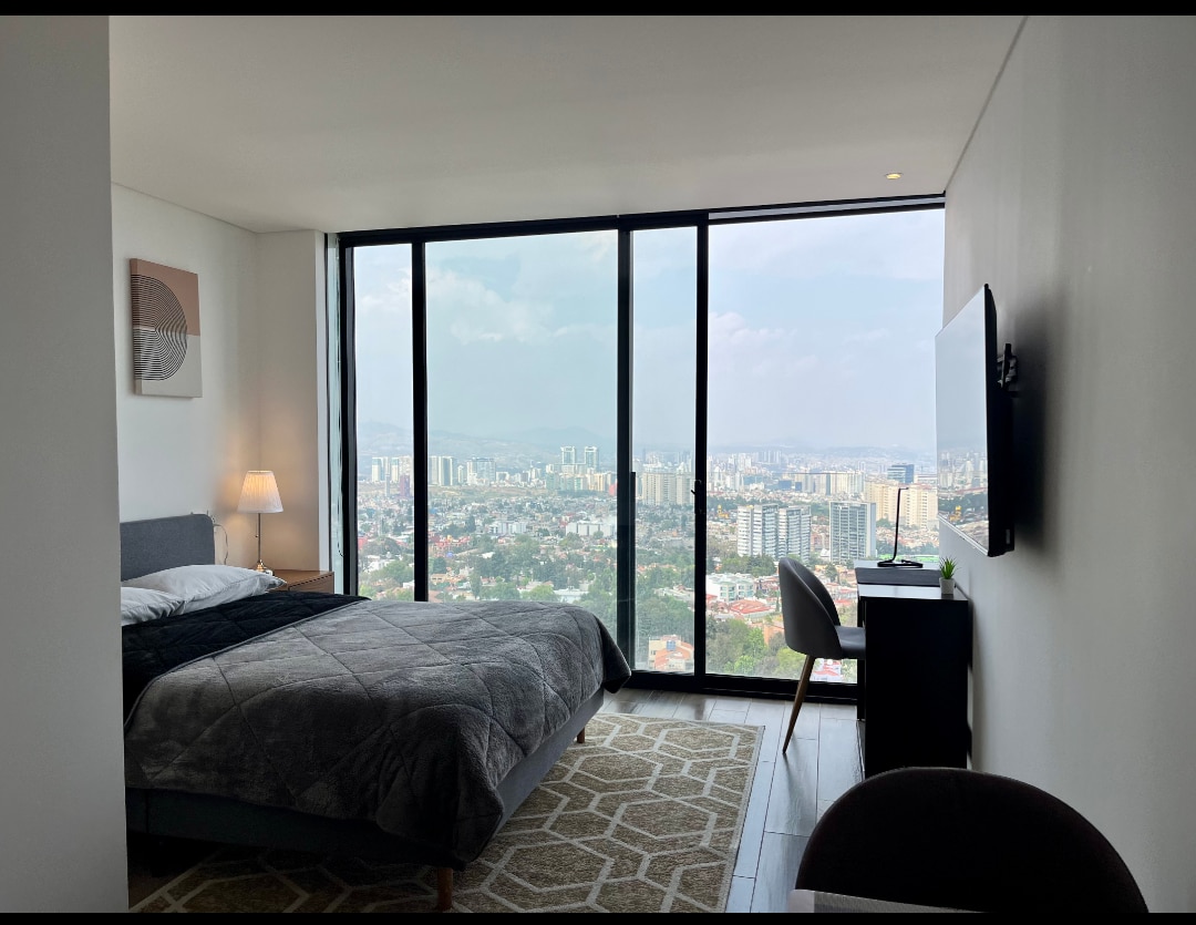 A modern bedroom features floor-to-ceiling windows offering expansive views of the city skyline. A spacious bed is draped with a dark gray blanket. A bedside lamp provides soft lighting, while a small desk and chair are positioned beside the window.