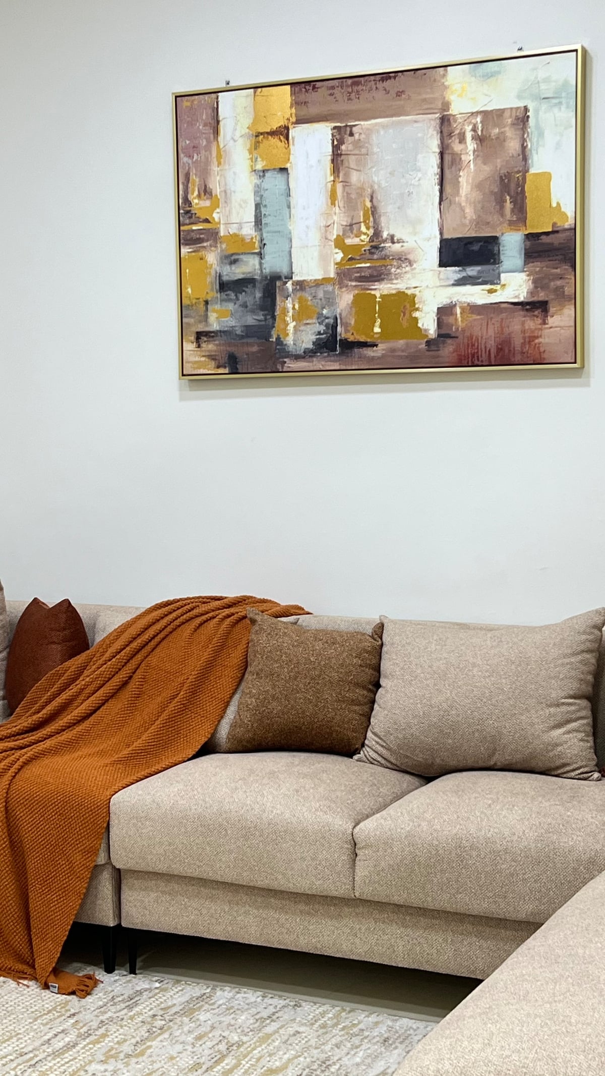 A comfortable sofa is displayed with warm-toned throw pillows and a textured orange blanket draped over it. Above the sofa, an abstract art piece featuring muted colors adds a decorative touch to the light-colored wall.
