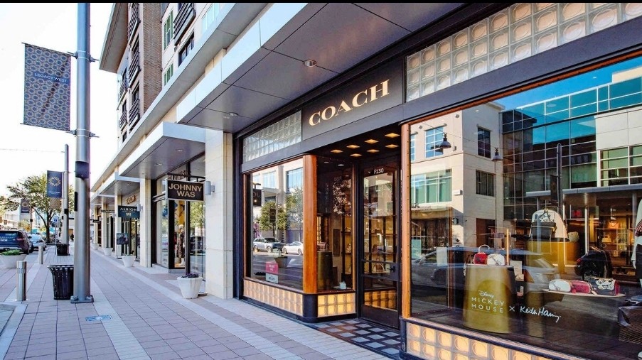 A stylish shopping street is showcased, featuring modern storefronts with large glass windows and vibrant displays. The Coach store is prominently seen, with its logo clearly visible. Pedestrian pathways are lined with decorative banners and well-maintained sidewalks, indicating a welcoming atmosphere.