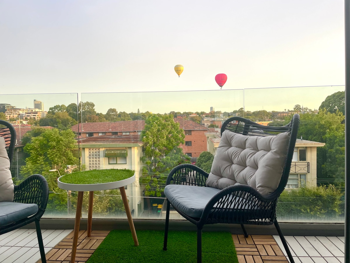 A balcony is showcased with two comfortable chairs and a small round table placed on synthetic grass. Scenic views of neighboring buildings and balloons floating in the sky enhance the outdoor space, providing a peaceful atmosphere.