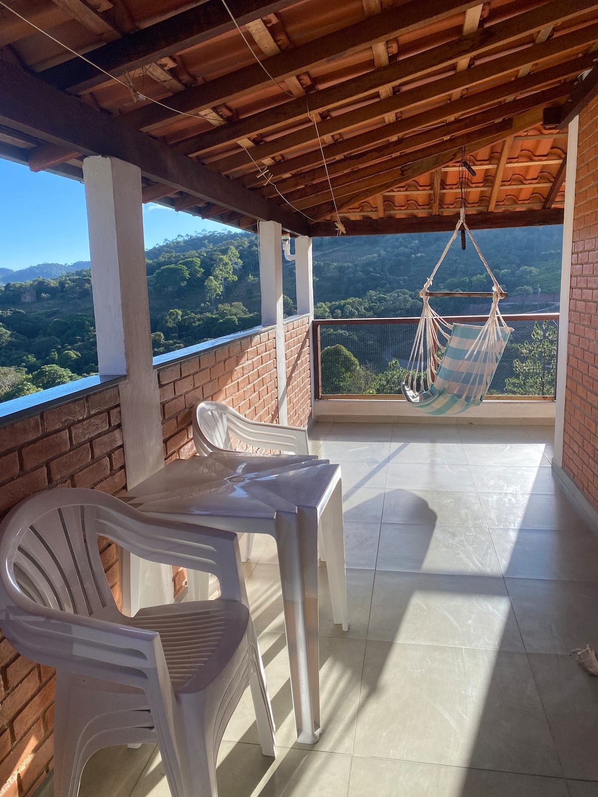 A balcony area is visible, featuring two white plastic chairs positioned on light-colored tiles. A hammock hangs from the ceiling, inviting relaxation. Natural light fills the space, casting soft shadows. The background showcases green hills under a clear blue sky.