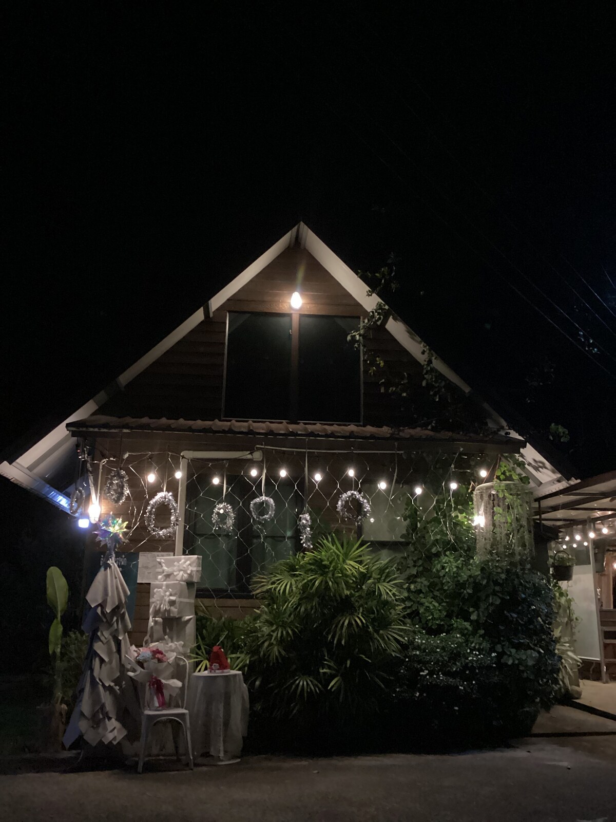 A charming A-frame structure is softly illuminated by string lights, outlining the entrance. Lush greenery surrounds the building, with a small table set beside the door. The large window above reflects the warm glow, inviting guests into the peaceful atmosphere.