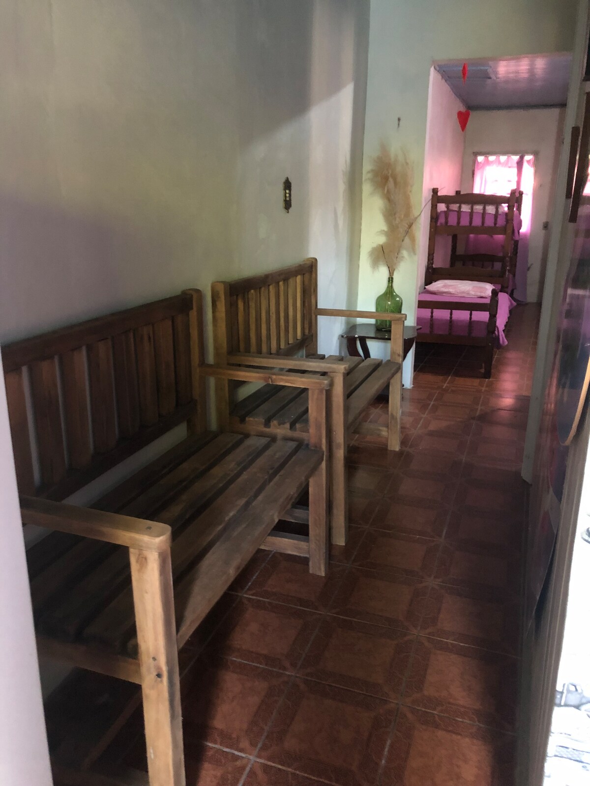 A narrow hallway features two wooden benches positioned against the wall. A subtle pink light fills the space, illuminating the warm wood tones. In the background, a small room containing a set of bunk beds is visible, enhancing the communal atmosphere.