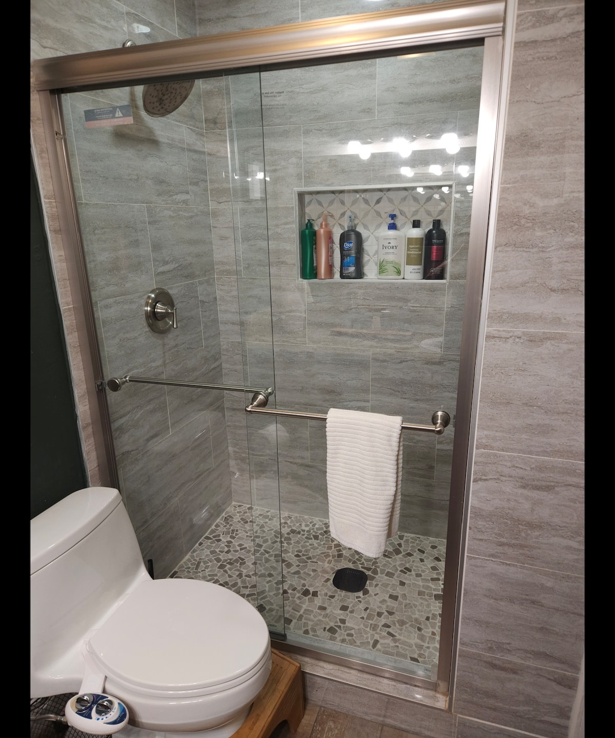 A modern shower area features a glass enclosure with a built-in niche displaying various toiletries. Decorative stone tiles cover the shower floor, while a white towel hangs neatly on a polished bar. A toilet is positioned adjacent to the shower, offering functionality.