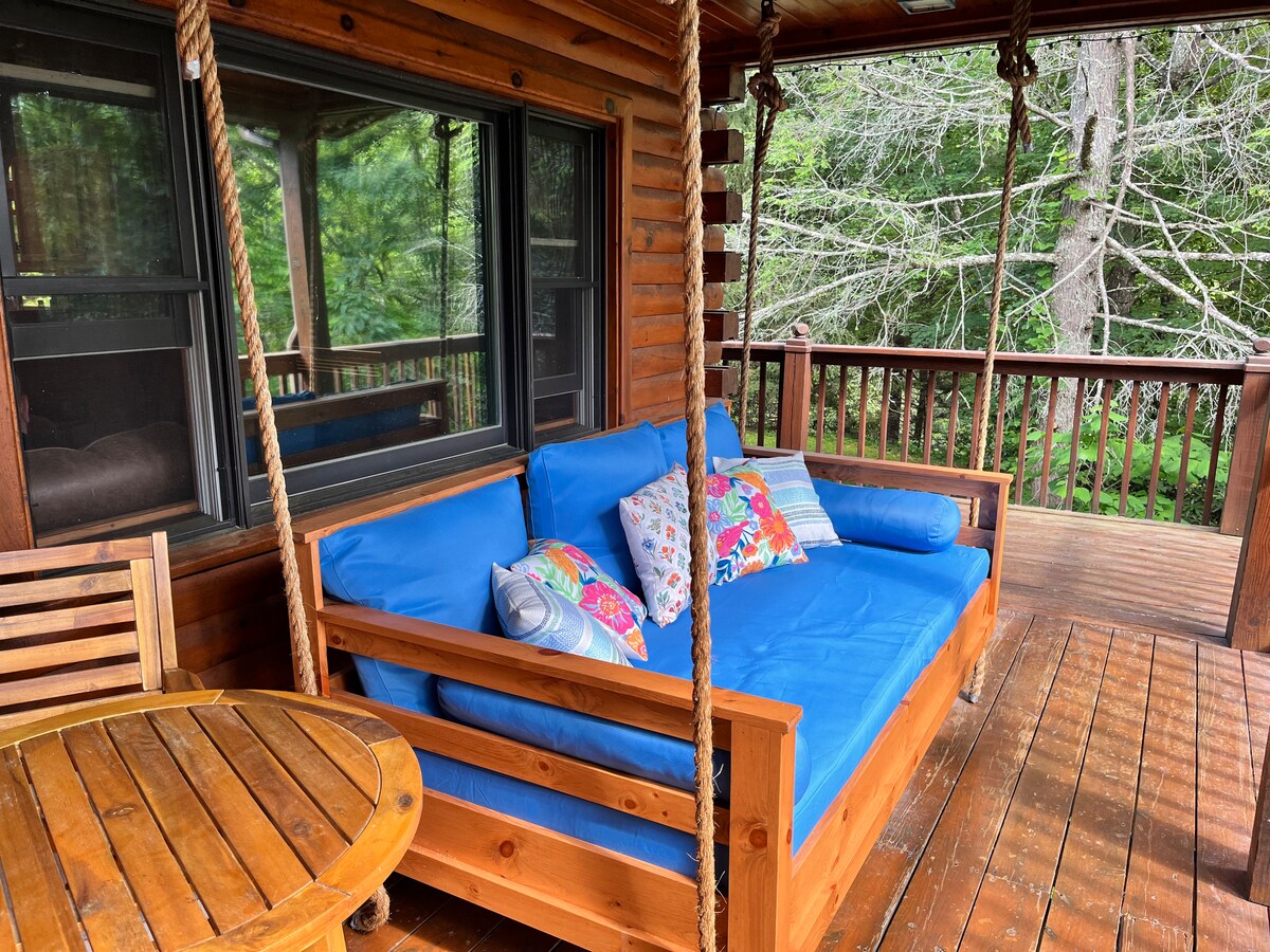 A shaded outdoor seating area is featured with a wooden frame and a blue cushioned sofa adorned with colorful throw pillows. A round wooden table is nearby, situated on a wooden deck that overlooks a serene natural setting.