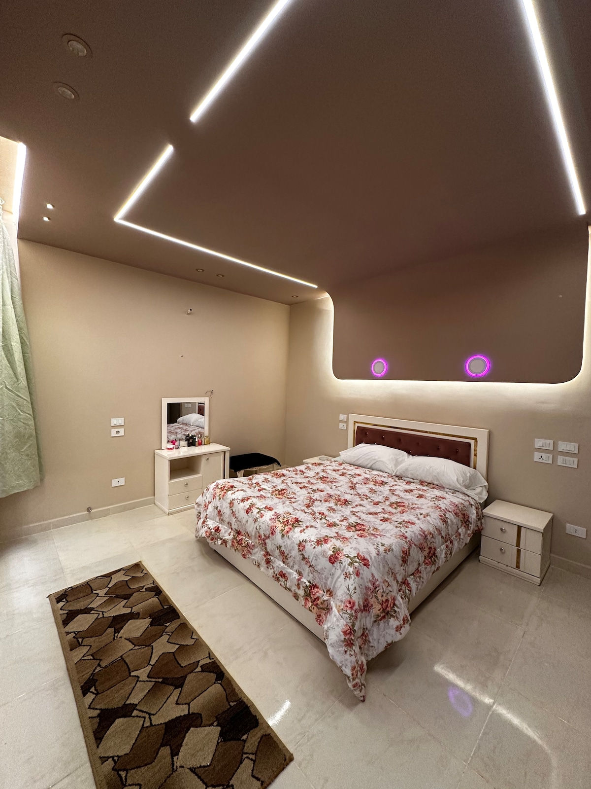 A bright and modern bedroom features a spacious bed dressed in floral bedding. Soft lighting is provided by illuminated ceiling panels, while a small bedside table and a dresser are positioned against the walls. A window with light green curtains allows natural light to enter the room.