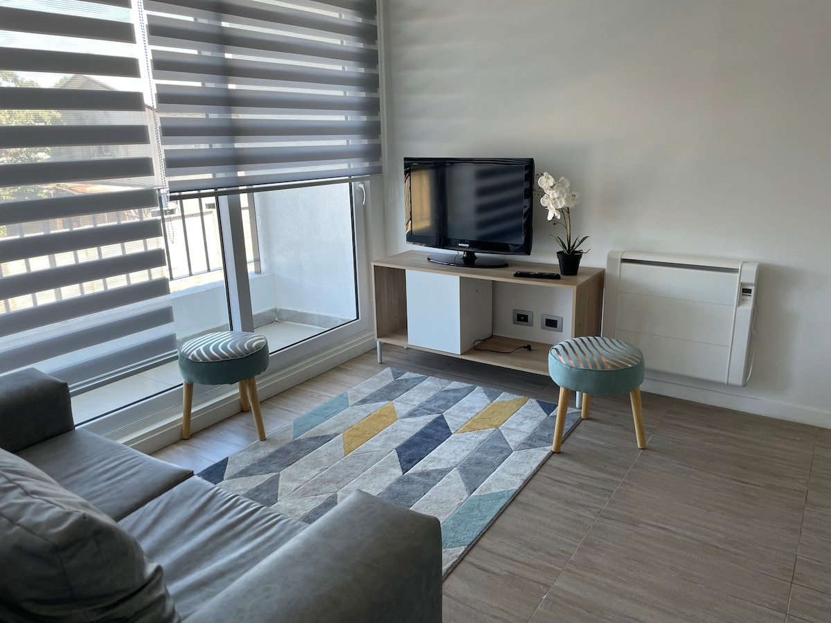A modern living area features a comfortable gray sofa paired with two small stools, positioned beside a geometric patterned rug. A television rests on a wooden stand, accompanied by a plant in a pot. Large windows with striped shades allow natural light to enter.