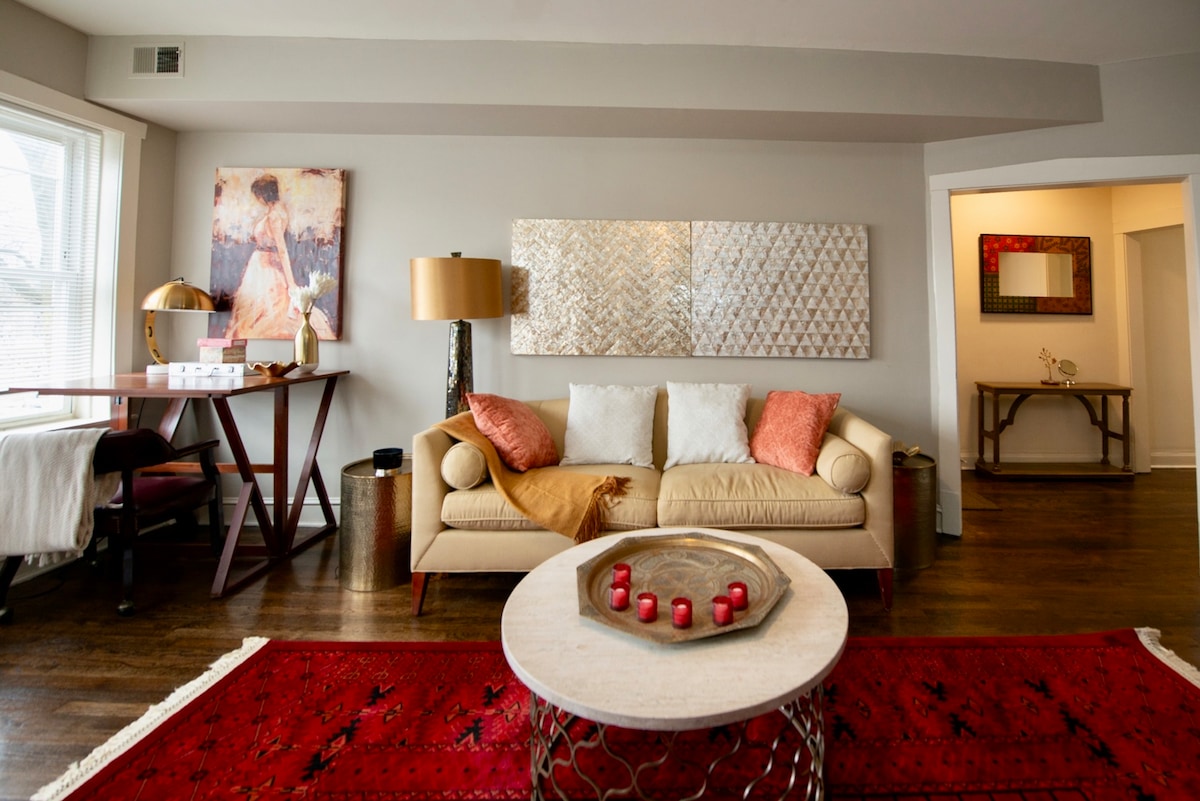 A spacious living area is presented with a neutral sofa adorned with decorative pillows. A round coffee table, surrounded by a vibrant red area rug, sits center stage. An artistic piece and a stylish floor lamp enhance the room's inviting ambiance, while a dining table is positioned nearby.