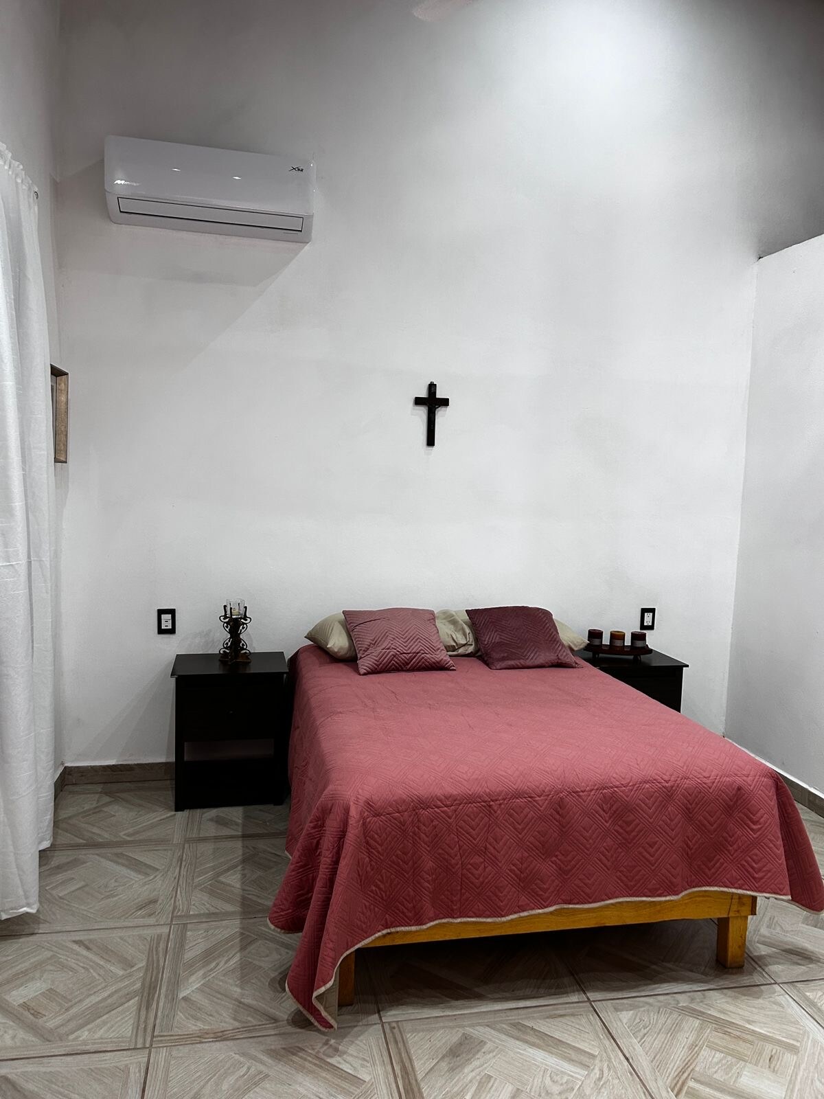 A cozy sleeping area features a double bed covered with a maroon blanket, accompanied by two decorative pillows. On either side of the bed, dark wood nightstands house a candle and a decorative item. A simple wooden cross is mounted on the light-colored wall.