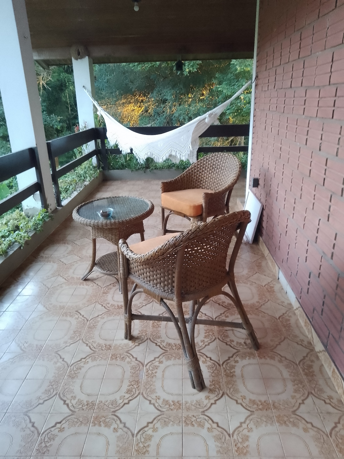 A welcoming porch is presented with two wicker chairs and a matching table. A hammock is gently draped in the background, surrounded by greenery. The tiled floor features an intricate design, complementing the wooden ceiling and the view of nature beyond the railing.