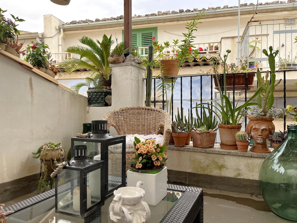 A private terrace is adorned with various potted plants and flowers. A wicker chair is positioned near a table, which holds decorative lanterns and a small flower arrangement. The surrounding area features greenery and a glimpse of neighboring buildings with open doors and windows.