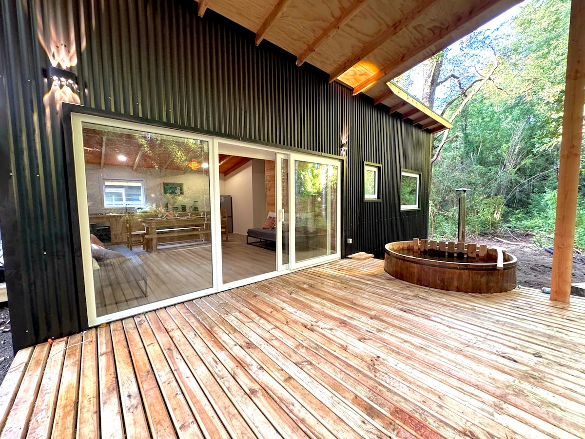 A cabin exterior is framed by natural surroundings, featuring a wooden deck that leads to a round wooden hot tub. Large glass doors offer views into a cozy interior, while two small windows illuminate the space. The design highlights a modern aesthetic combined with natural materials.