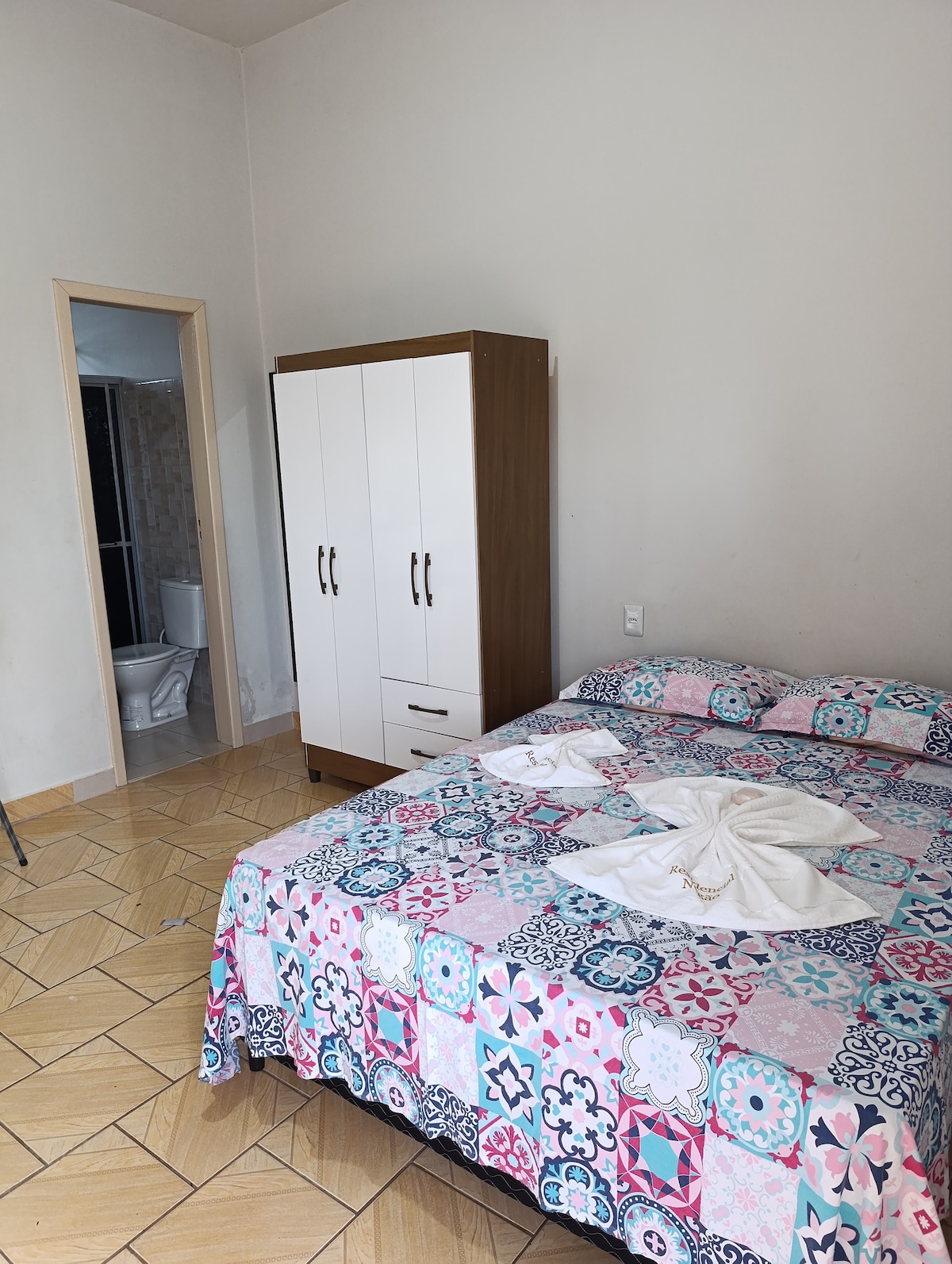 A sleeping area is designed with a colorful patterned bedspread covering a spacious bed. A white wardrobe stands against the wall near a doorway, leading to a separate bathroom. Light-colored tiles cover the floor, enhancing the room's bright atmosphere.