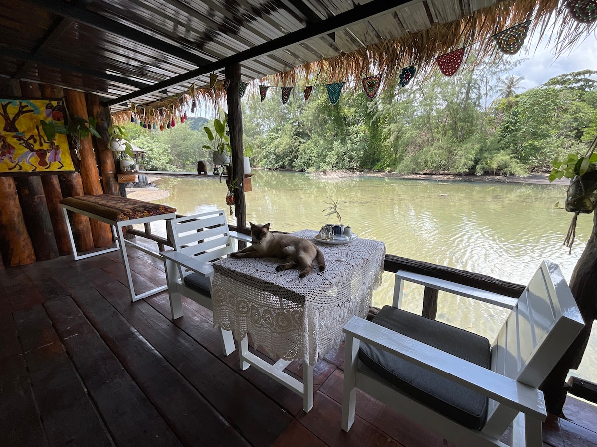 An outdoor seating area is situated along the water, featuring a white table and chairs under a thatched roof. A cat rests peacefully on the table, while greenery frames the view of the calm river.