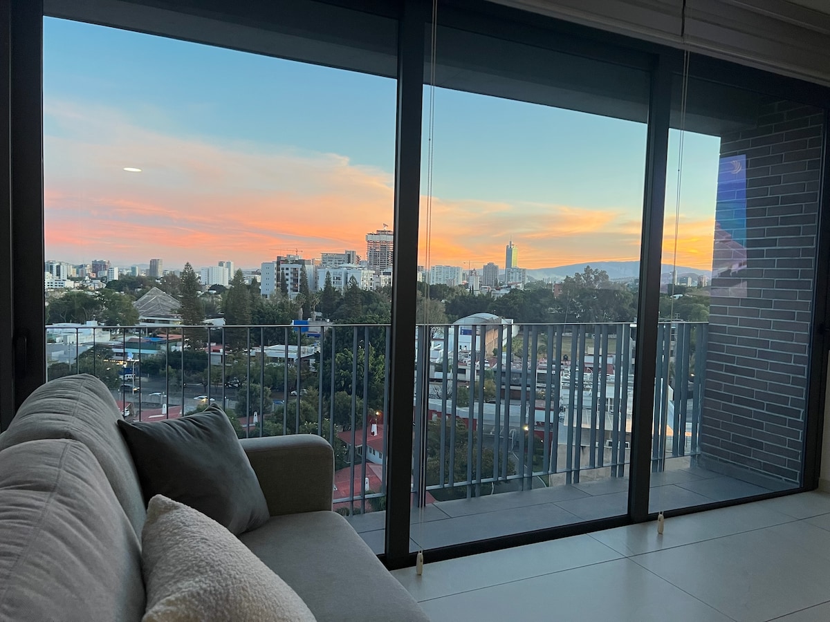 A spacious living area features a large window providing a panoramic view of the city skyline at sunset. Soft hues of orange and pink illuminate the sky, while a comfortable sofa is positioned nearby, inviting relaxation and enjoyment of the scenic landscape.