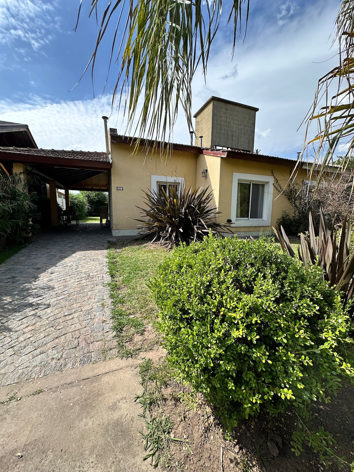 The exterior of a single-story house is visible, featuring a light yellow facade and a chimney. A pathway made of cobblestones leads towards the entrance, surrounded by various shrubs and plants. A clear blue sky and soft clouds are observed above.