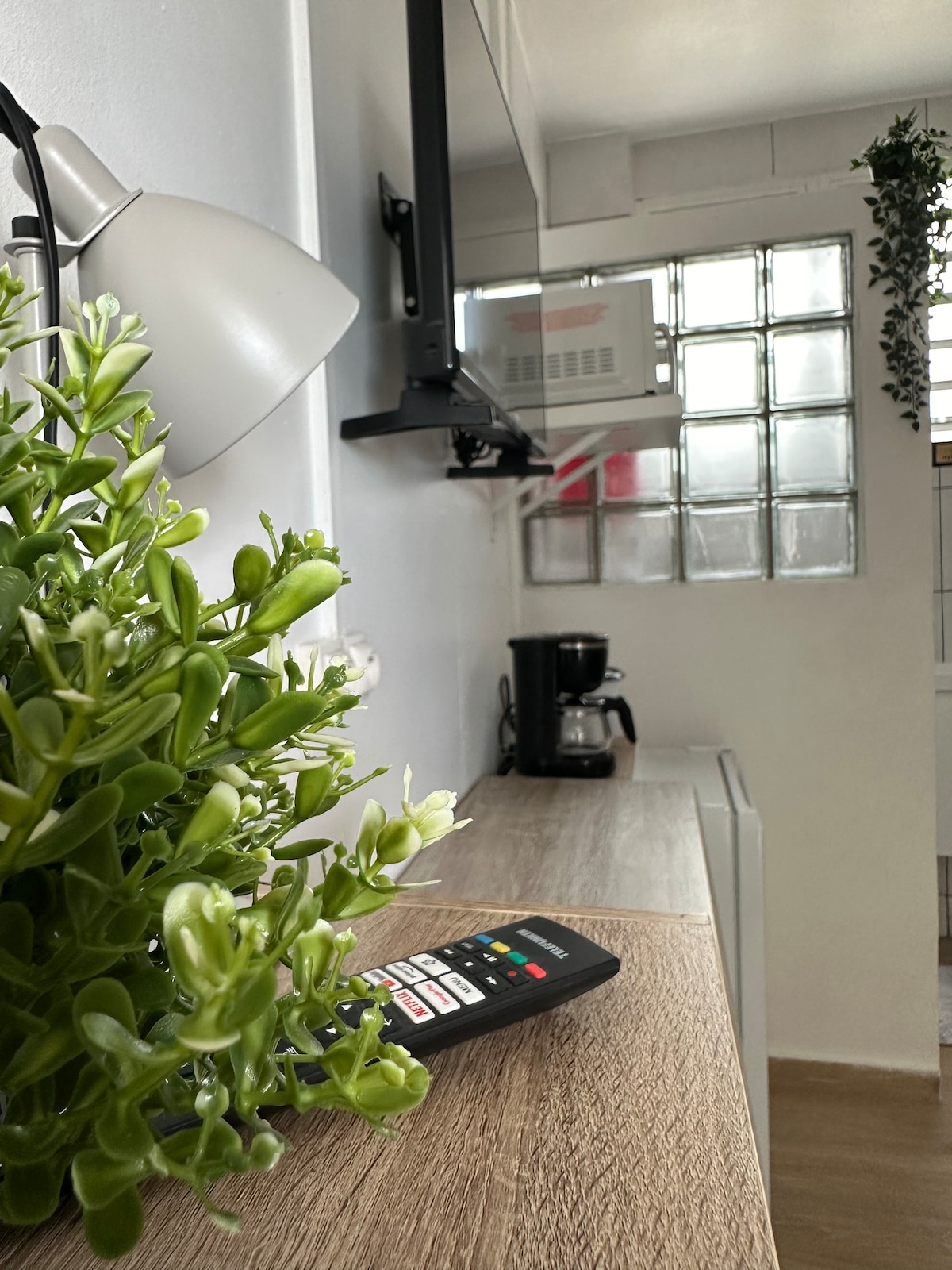 A clean and functional kitchenette is visible, featuring a modern coffee maker, microwave, and refrigerator. A small potted plant adds a touch of greenery. The space is illuminated by a sleek lamp positioned on the countertop, complementing the light color scheme.