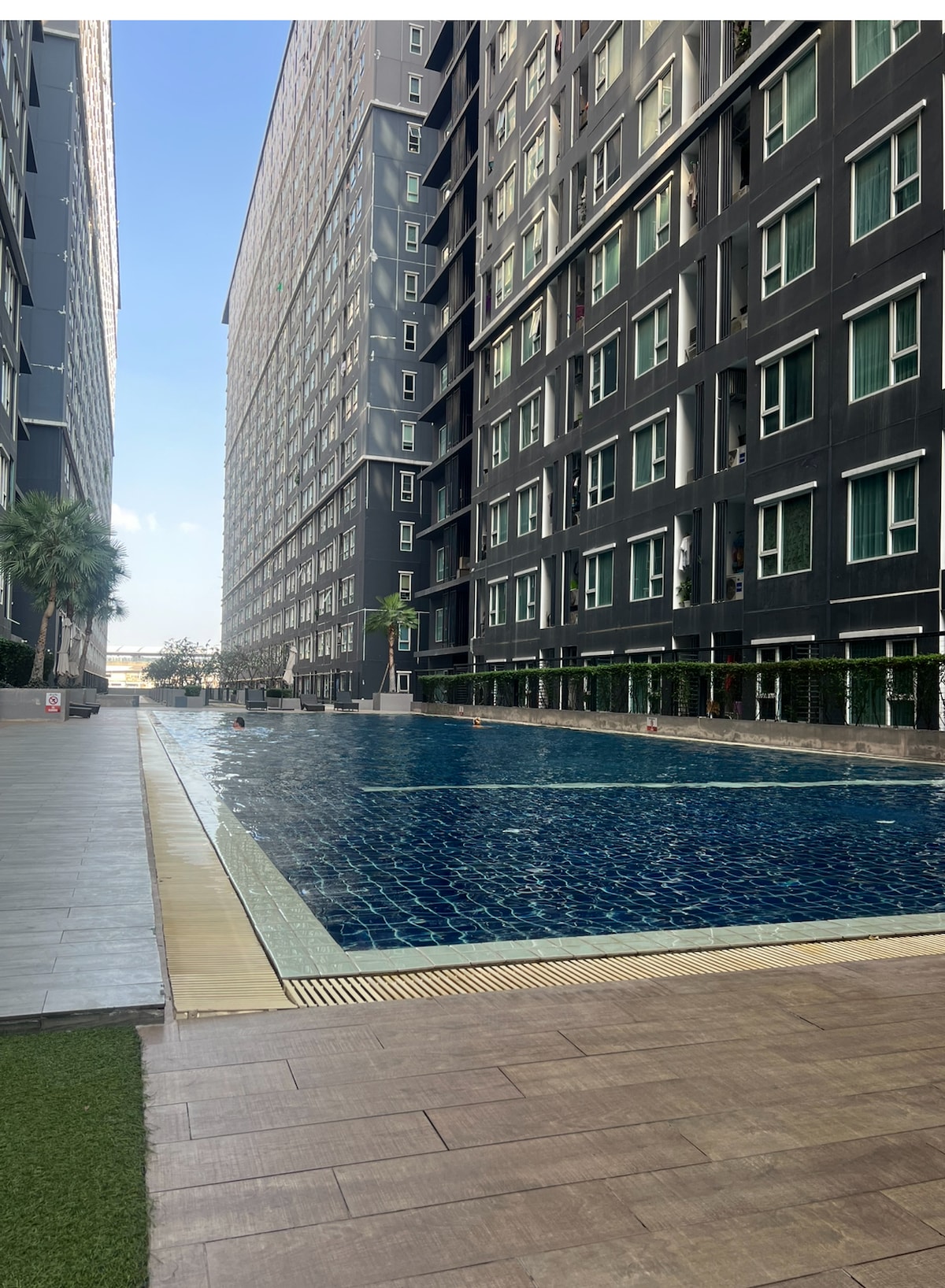 A serene swimming pool is situated between two modern apartment buildings, with clear blue water inviting visitors. Lush greenery frames the pool area, and the sky above is bright, enhancing the peaceful ambiance. The pool's tiled surface reflects the surrounding structures.