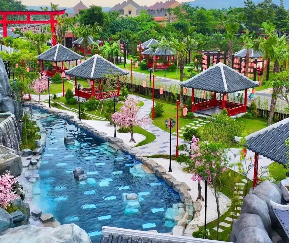 A serene outdoor space is presented, featuring a winding pool surrounded by lush greenery and landscaped gardens. Traditional-style pavilions are scattered throughout, with cherry blossom trees adding subtle color. Rocky formations and a gentle waterfall create a tranquil backdrop, enhancing the peaceful ambiance.
