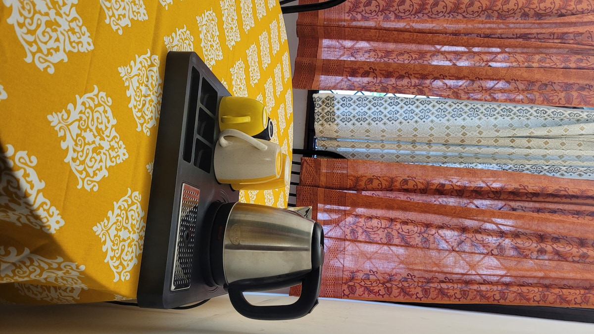 A kitchen area features a bright yellow tablecloth adorned with a white pattern. A metallic kettle sits on a tray beside two cups, one of which is yellow and the other white. Sunlight filters through semi-transparent curtains, creating a warm ambiance.
