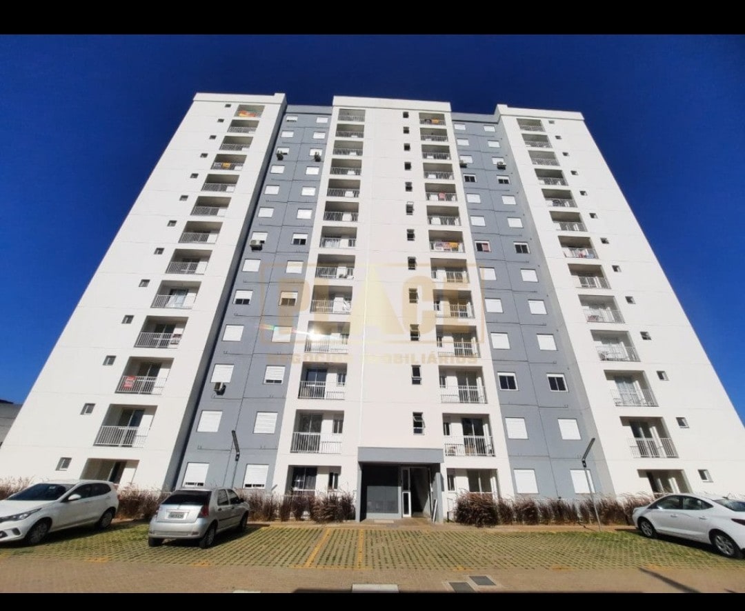 A modern multi-story building is depicted with a mix of white and gray façades. A clear blue sky serves as the backdrop. The ground level features an entrance with covered access, while vehicles are parked in the foreground on a paved surface.